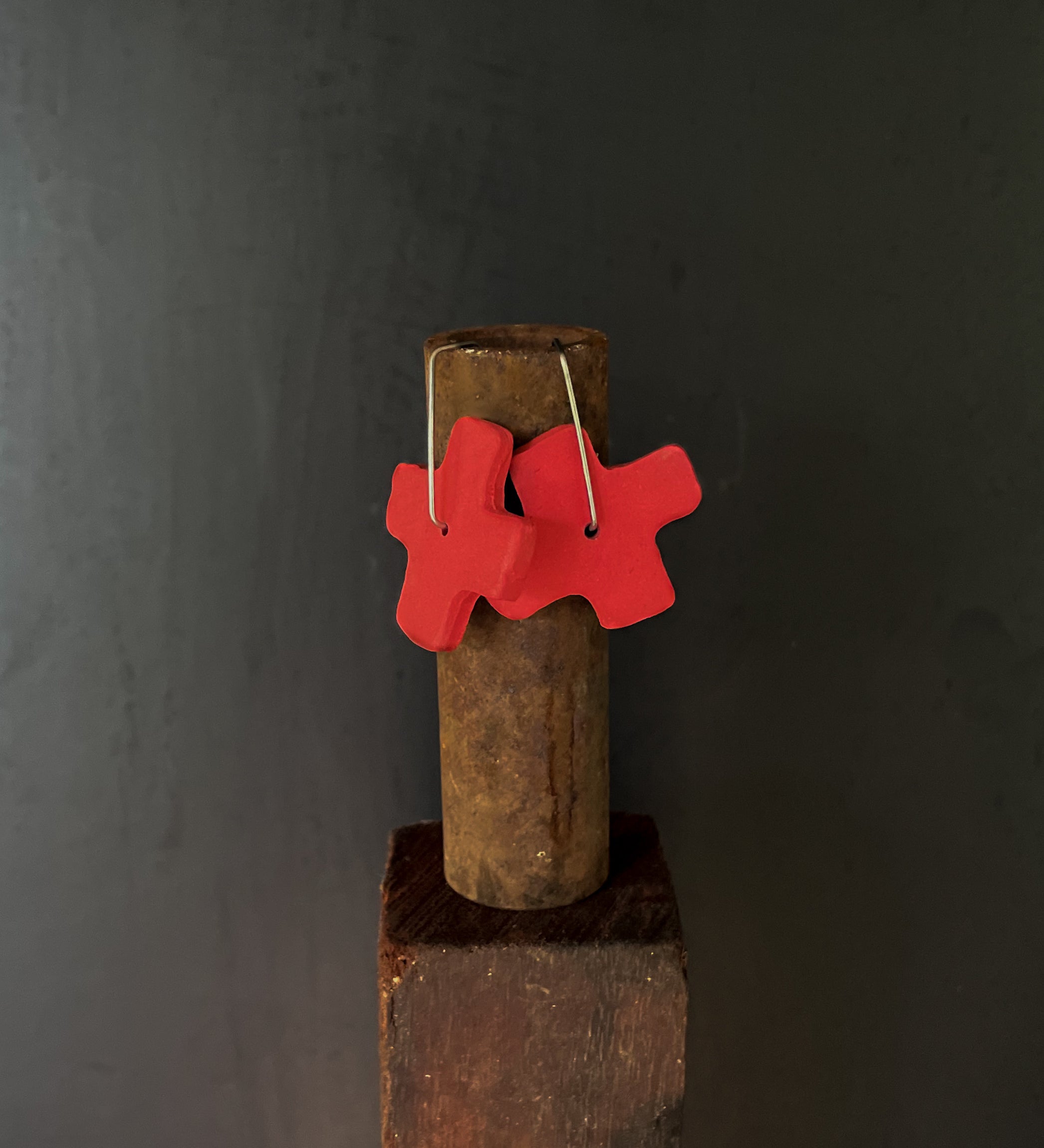 Red x-shaped earrings hanging on a rusty metal rod against a dark background