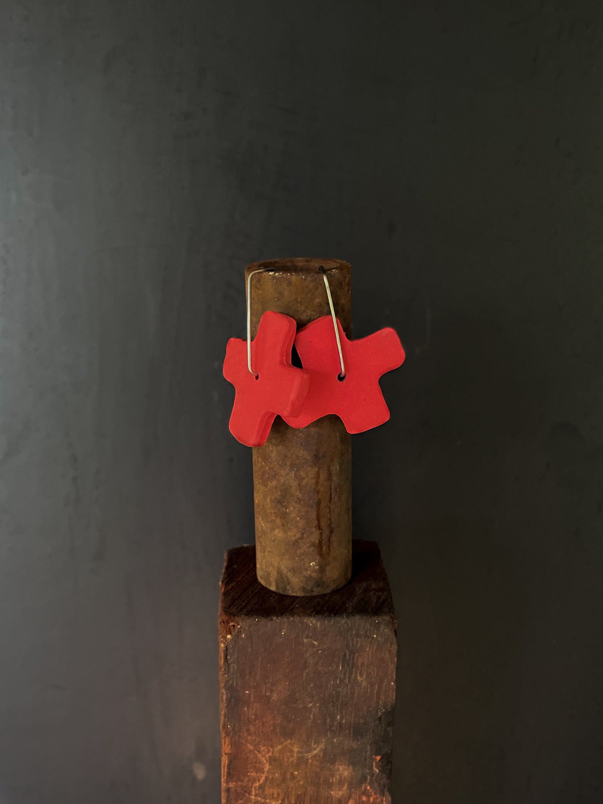 Red x-shaped earrings hanging on a rusty metal rod against a dark background