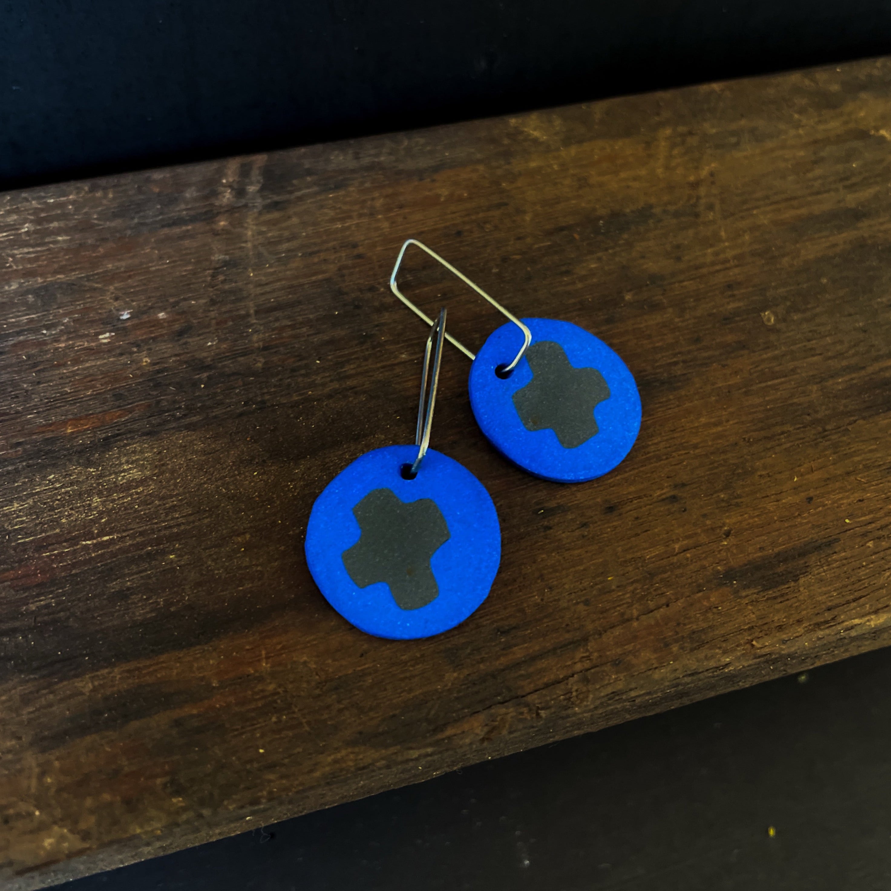 Blue circular earrings with a central black x design on a wooden surface.